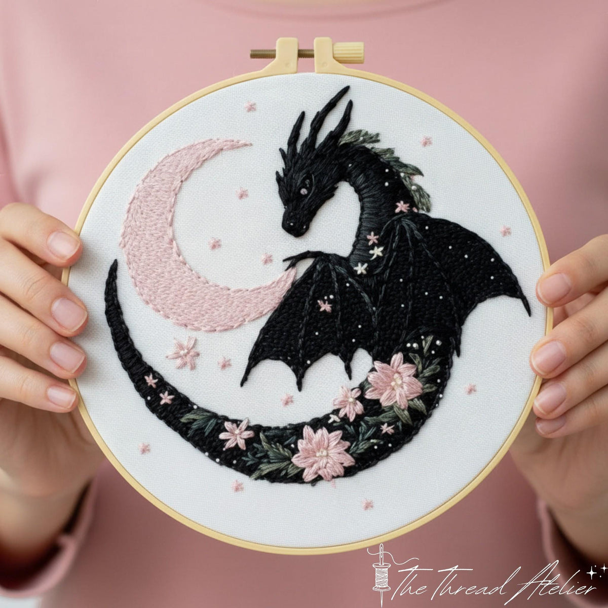 Black dragon and pink crescent moon stitched in a hoop, held against a pink jumper